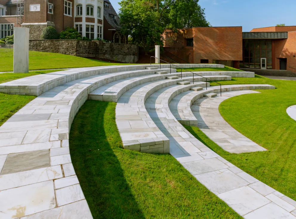 Tiered stone seating in a grassy outdoor amphitheater, with brick buildings and trees in the background under a clear blue sky.