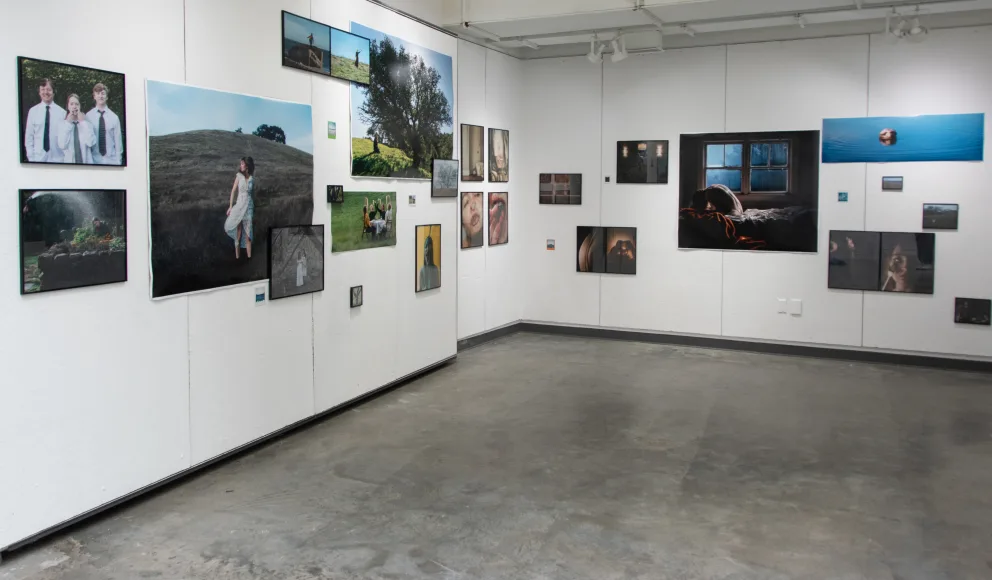 Art gallery with multiple photographs on white walls, including portraits and landscapes. Empty floor space in the center.