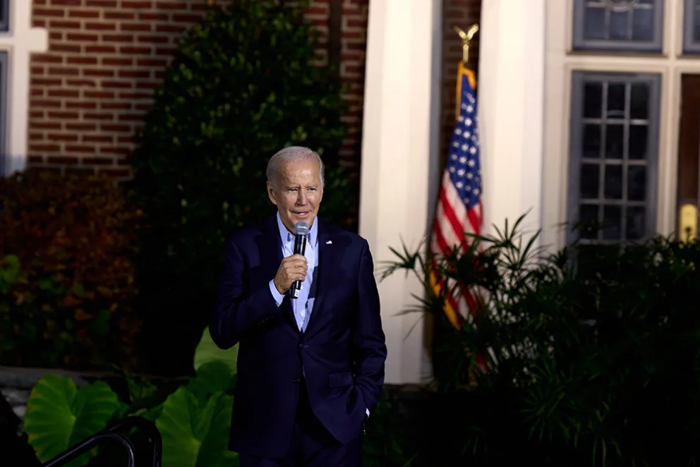 Individual in a suit holding a microphone stands outdoors, with an American flag and foliage in the background.