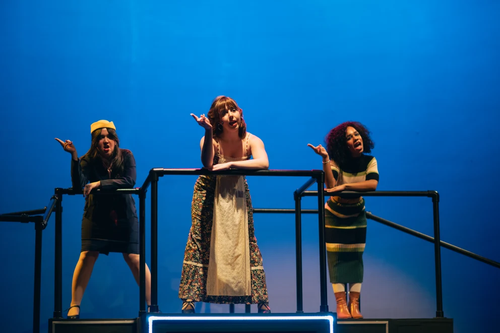 Three performers stand on a stage, each expressing with gestures and facial expressions, against a blue background.