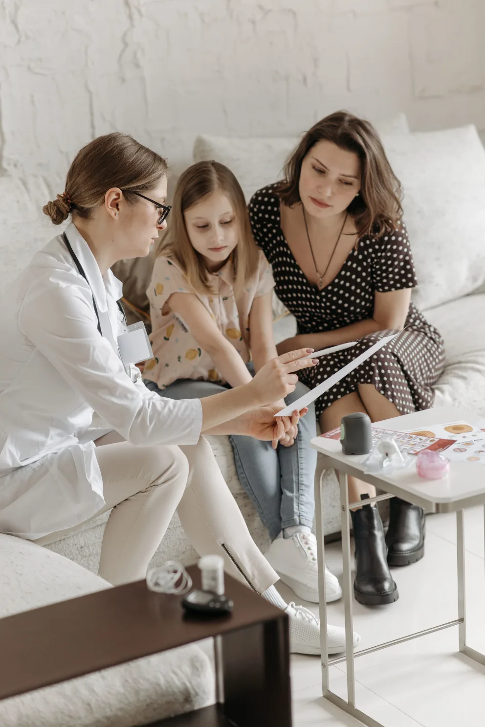 Person in a white coat showing papers to two seated individuals on a couch, with medical items on a table nearby.