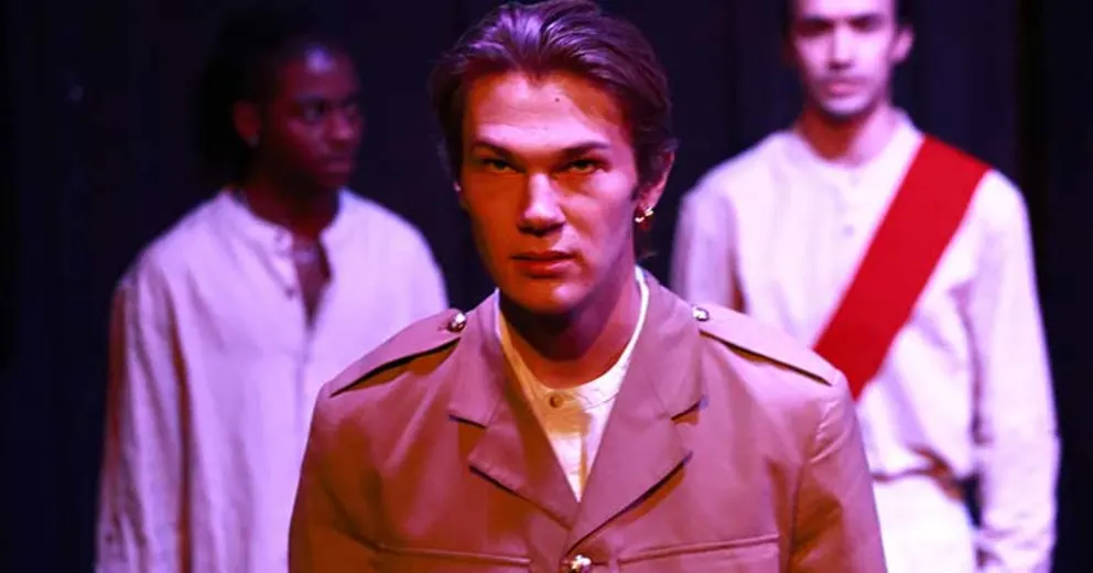 Person in a military-style jacket stands in focus, with two others in light clothing blurred in the background on a dimly lit stage.