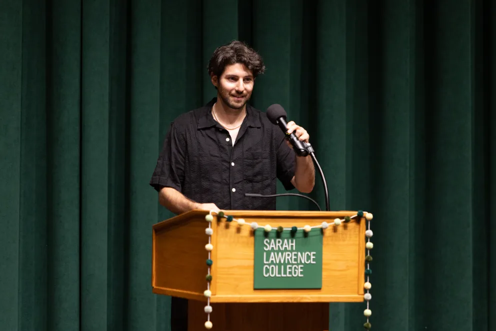 Person speaking at a podium with a microphone at Sarah Lawrence College, in front of green curtains.