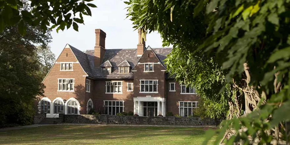 Large brick mansion with multiple chimneys, set in a leafy landscape, viewed through tree branches.