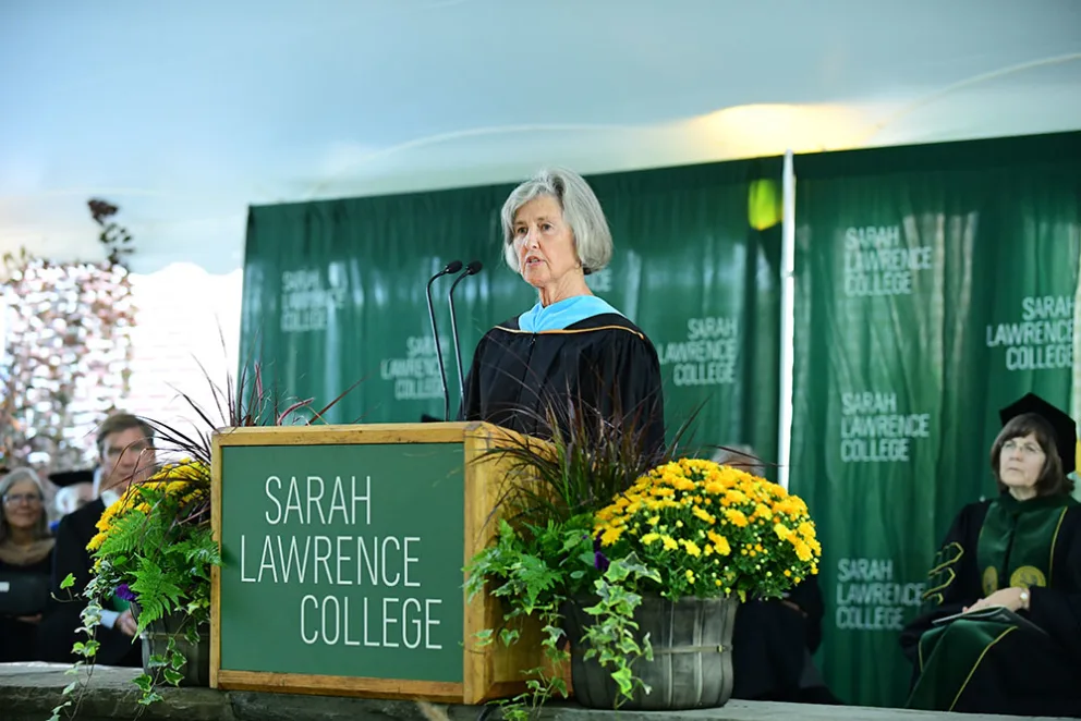 Person speaking at a podium with the Sarah Lawrence College logo, surrounded by plants and yellow flowers, in front of a green backdrop with audience seated.
