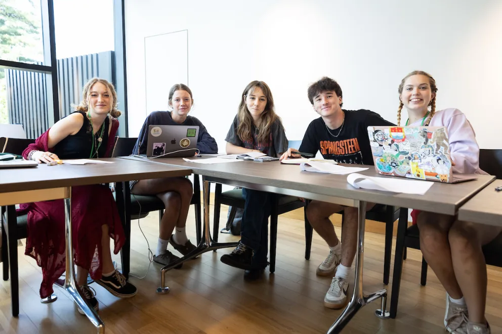 Five people sitting at a table in a classroom, with laptops and papers in front of them, smiling at the camera.