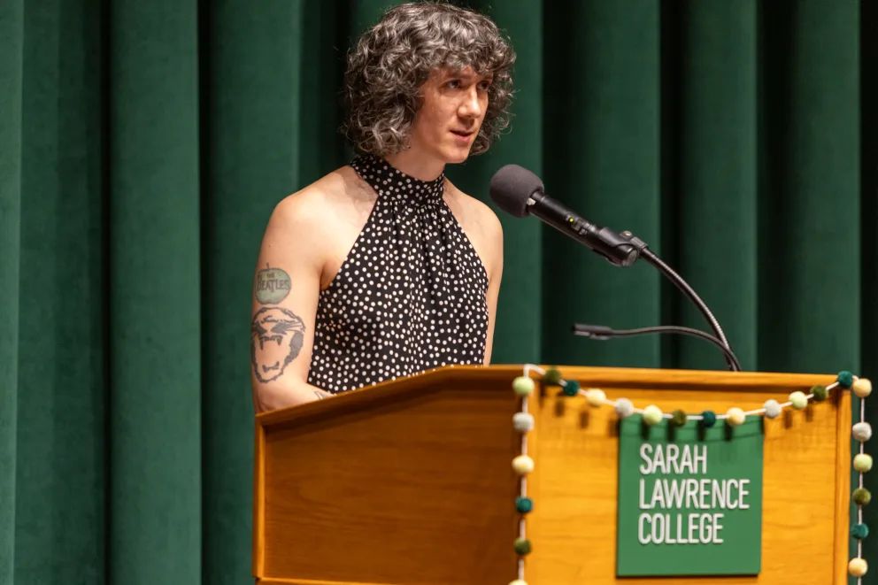 Person with curly hair and a tattoo speaks at a podium labeled Sarah Lawrence College, wearing a polka dot top.