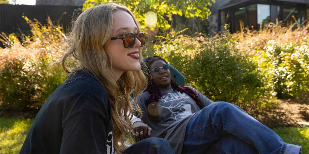 Two people sit on grass, enjoying sunlight. One leans back casually, the other sits upright wearing sunglasses. Bushes and trees in the background.