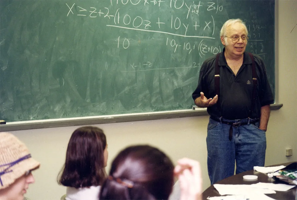 Person standing by a chalkboard with mathematical equations, gesturing while teaching a group of individuals in a classroom.