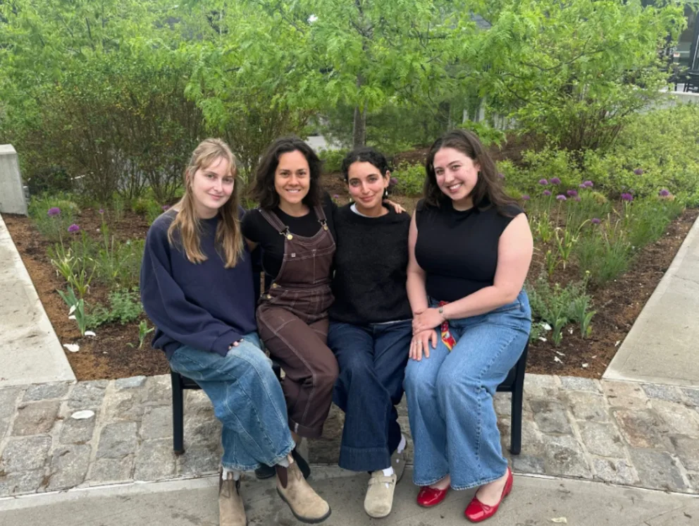Four people sitting on a bench outdoors, smiling, with a green, leafy background.