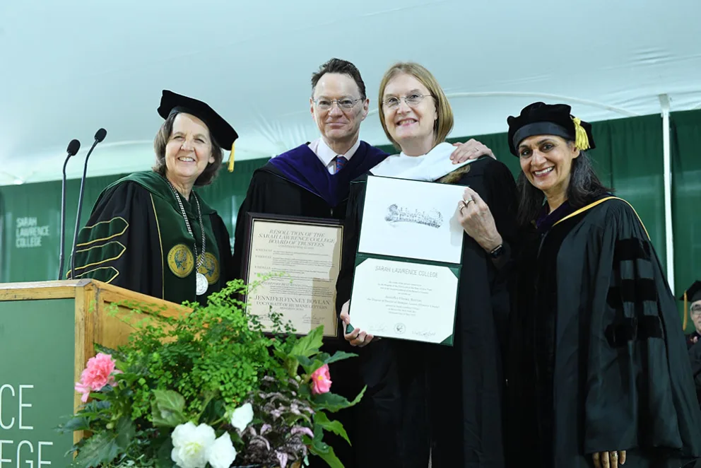Four people in academic regalia stand together at a podium, holding certificates. A floral arrangement is in the foreground, with a green backdrop.