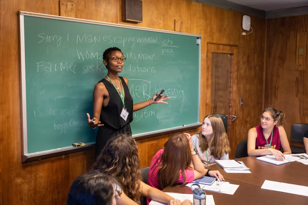 Person speaking to a group of attentive individuals seated around a table with notebooks in front of a chalkboard.