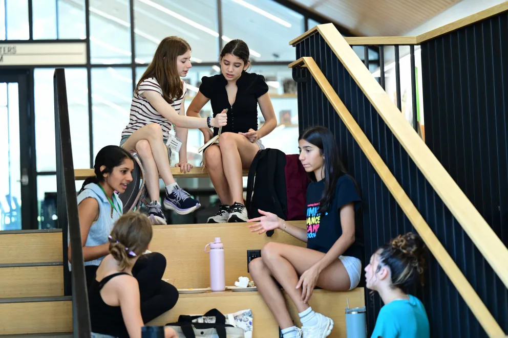 People sit and chat on indoor stairs, surrounded by bags and water bottles, in a bright space with large windows.