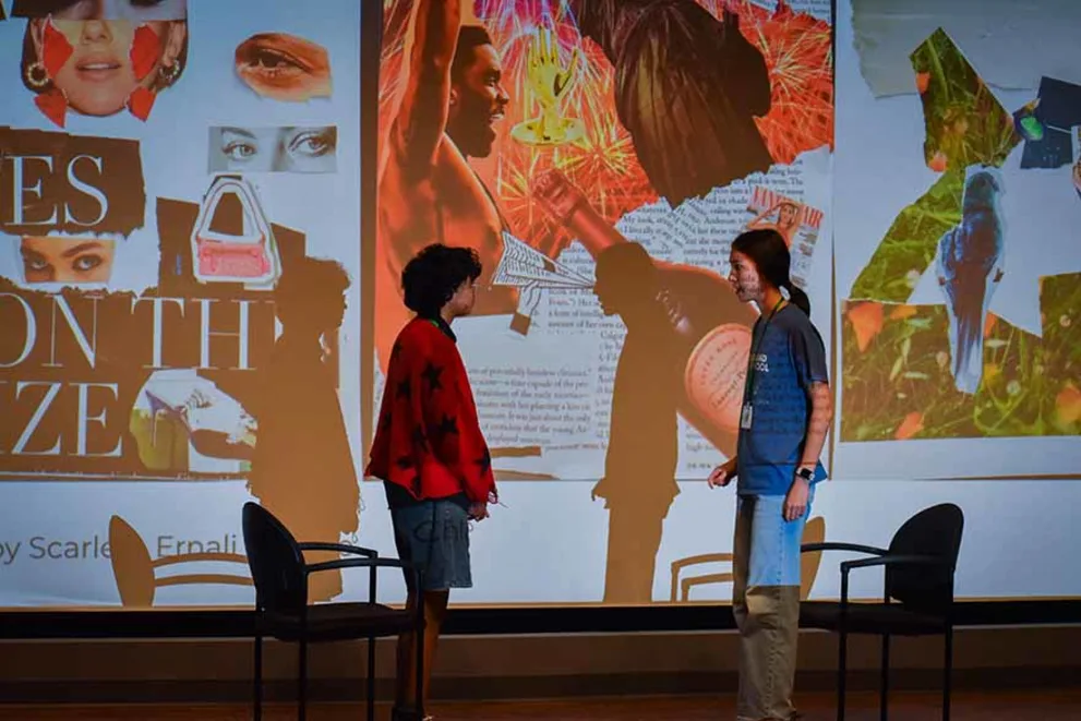 Two people stand facing each other on stage with chairs nearby, in front of a large colorful collage projection.
