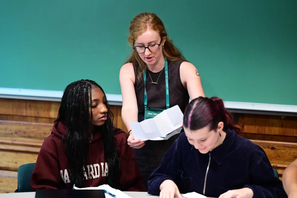 Person with glasses shows a paper to two others sitting at a table in a classroom with a chalkboard.