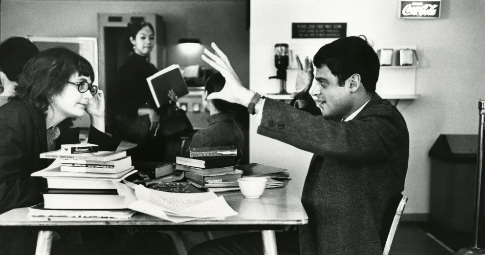 Two people at a table with books engaged in animated conversation. Another person walks in the background holding a book.