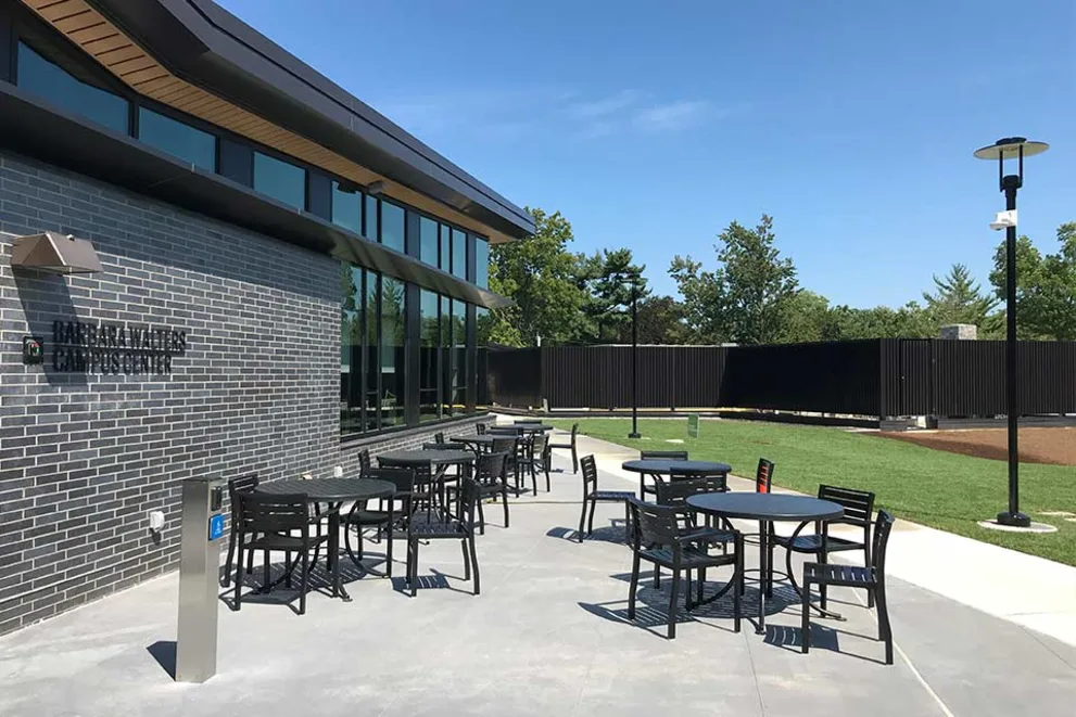 Outdoor patio with round tables and chairs next to a brick building under a clear blue sky, surrounded by grass and trees.