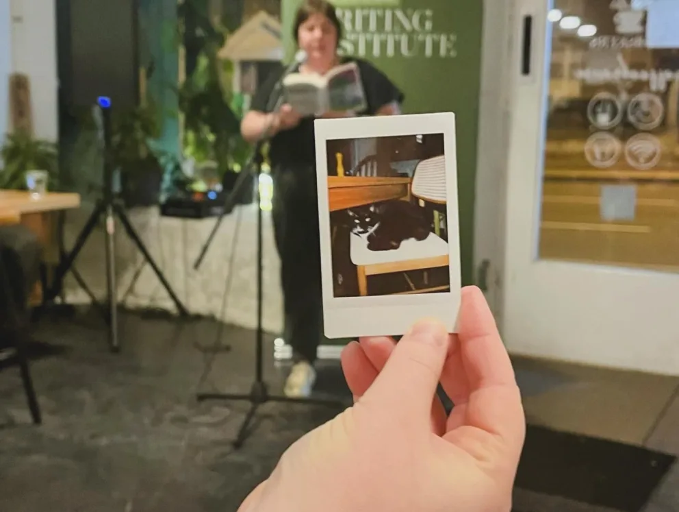 A person holding a Polaroid photo of a cat with someone reading in the background.