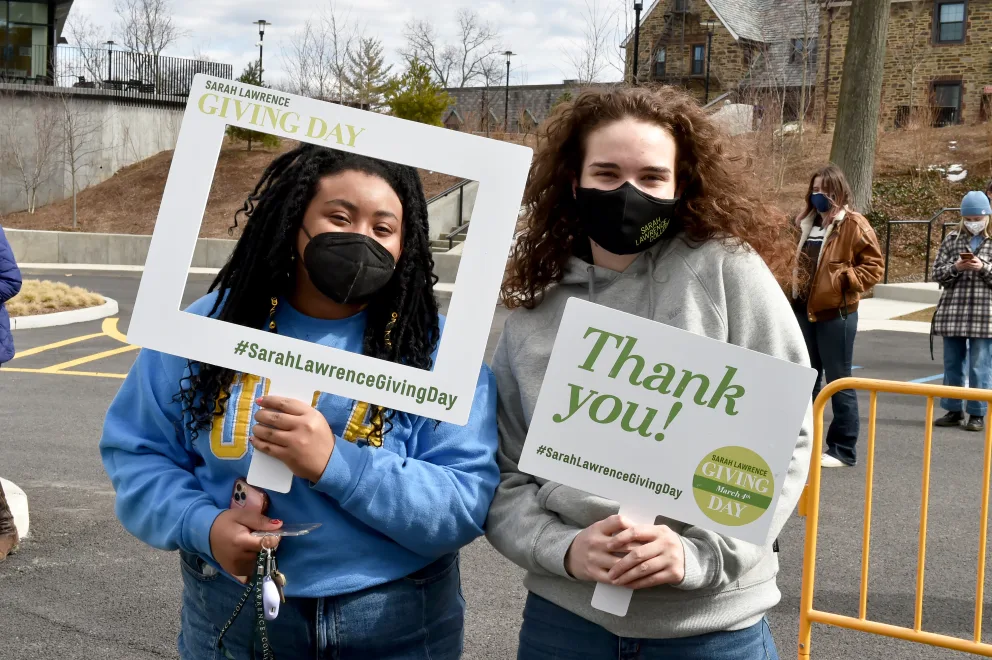 Two people outdoors holding signs for Sarah Lawrence Giving Day. One holds a frame, the other a "Thank you!" sign. Both wear masks.