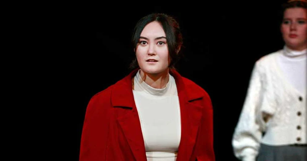 Person in a red coat looks ahead with a serious expression on stage, while another individual stands in the background wearing a white top.