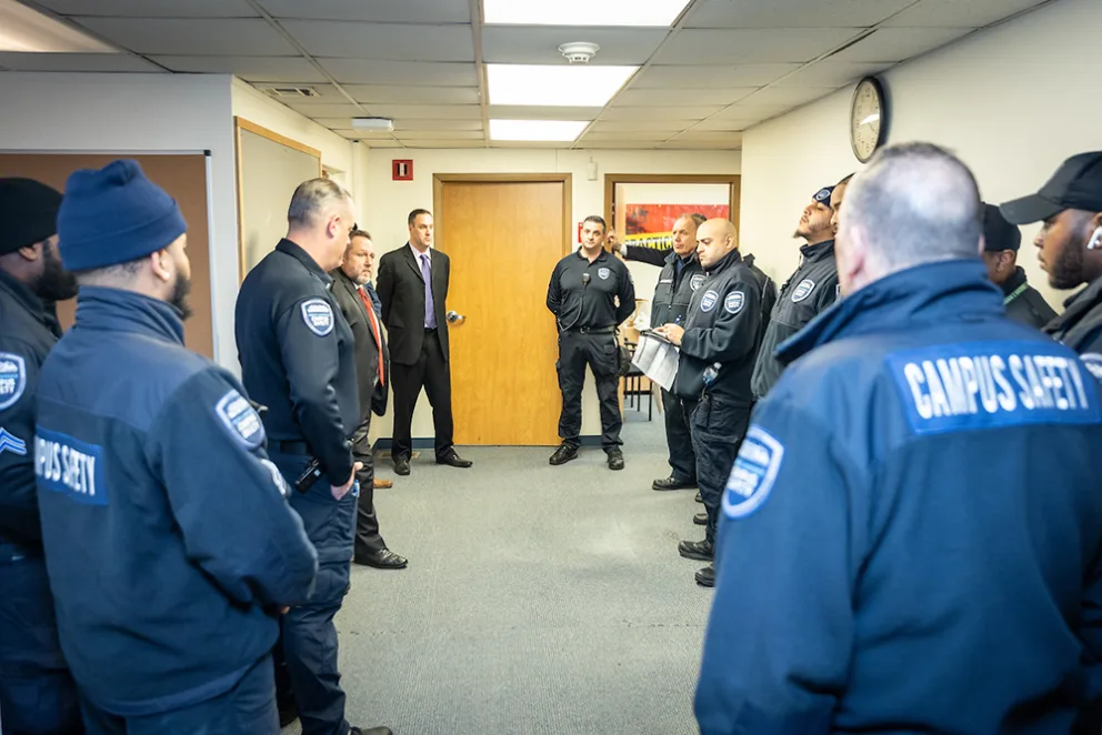 Group of campus safety officers in a meeting. Some wear uniforms with "Campus Safety" on jackets. Discussion taking place in a small room with a clock.