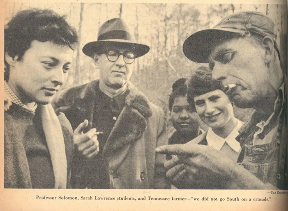 A group stands in conversation, one with a cigarette, in an outdoor setting; three wear coats and one overalls with a cap.