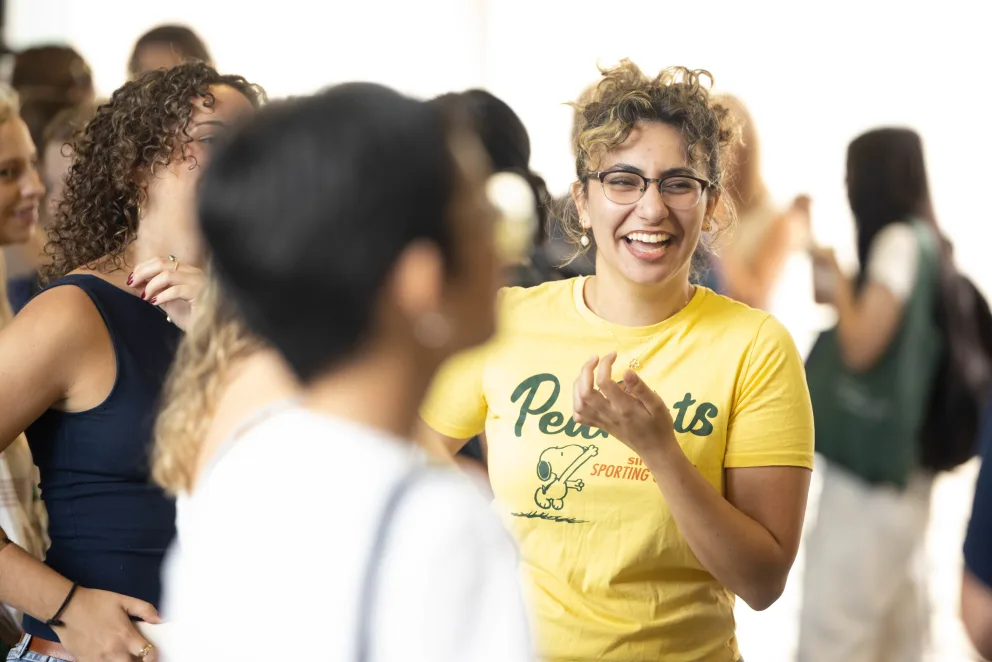 Person in yellow shirt and glasses smiles among a group in casual conversation. Bright setting with several people in the background.