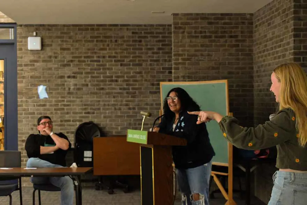 Person speaking at a podium smiles as someone points and another person watches in a room with brick walls.