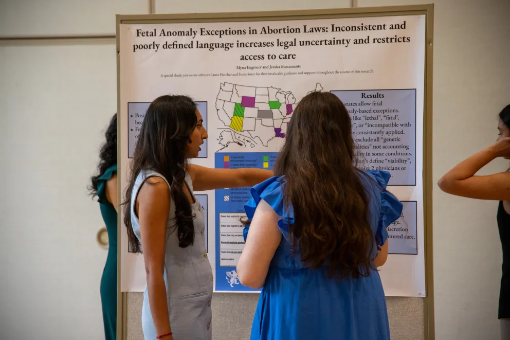 Two people discuss a research poster about fetal anomaly exceptions in abortion laws. One points to a U.S. map with highlighted states.