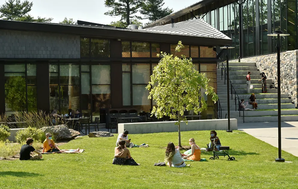 People sit on a grassy area reading and talking near a modern building with large windows and stone steps.
