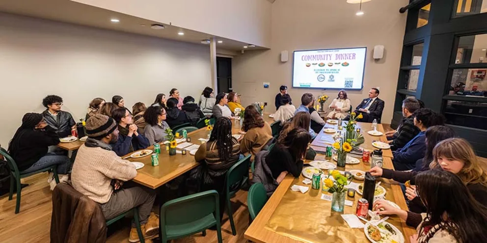 People seated around tables at a community dinner event. A panel speaks at the front under a "Community Dinner" screen.