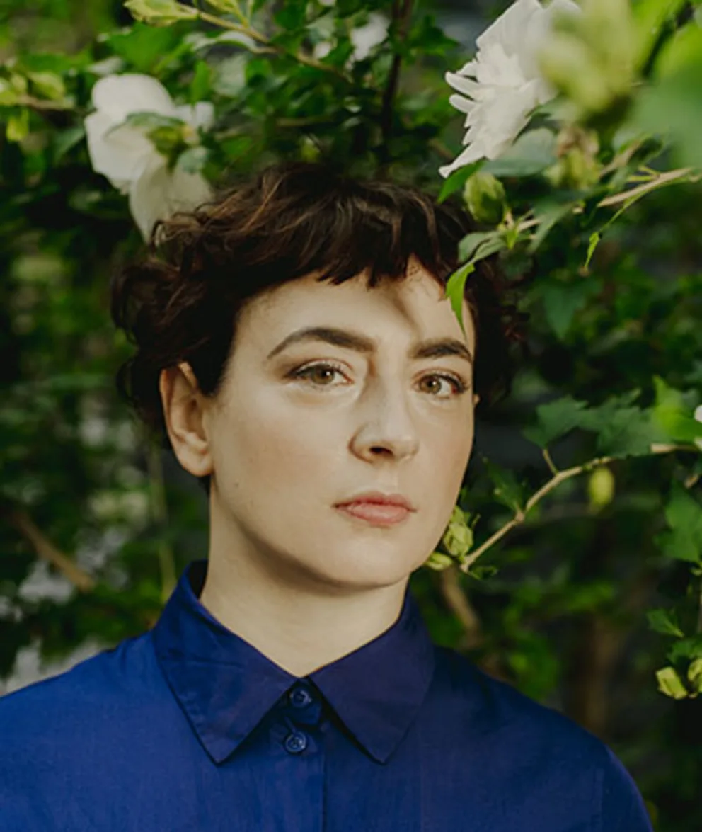 A person with short brown hair in a blue shirt stands in front of a bush with white flowers.