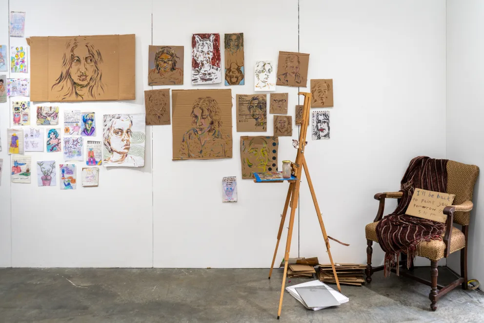 Art studio with various face sketches on walls, an easel, and a chair with a sign.
