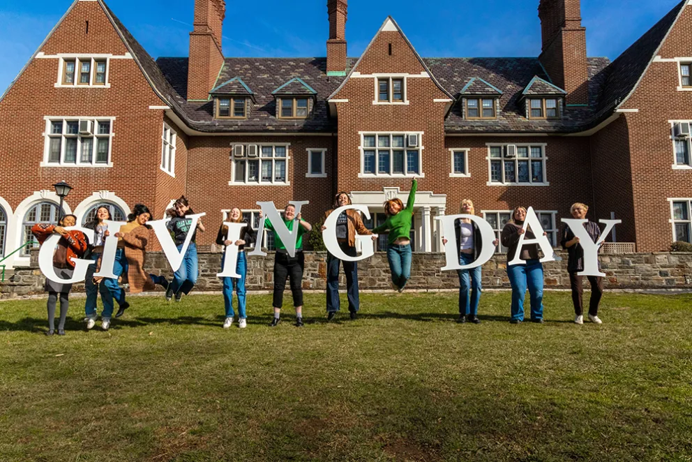 People holding large letters spelling "Giving Day" while jumping in front of a brick building.