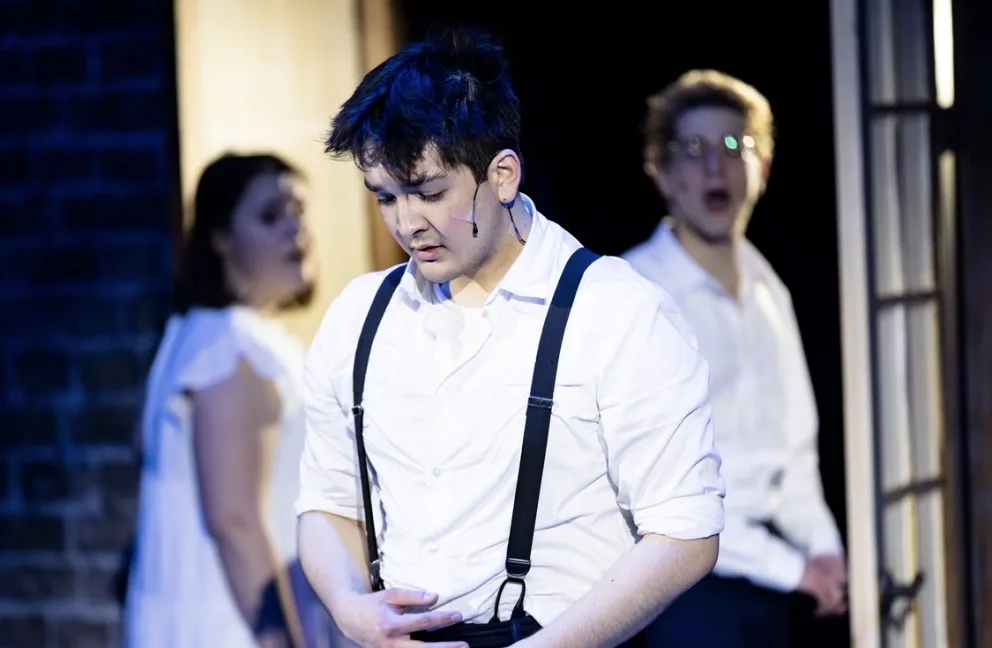 Person in a white shirt and suspenders performs on stage; two other performers in the background wearing white shirts.