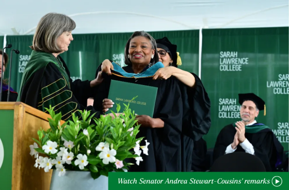 Graduate receives a diploma on stage at Sarah Lawrence College ceremony, surrounded by green banners and white flowers, with people applauding.