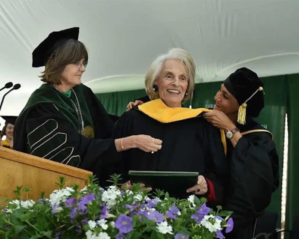 Three people in graduation attire, one receiving a diploma and yellow stole. A podium and flowers are in the foreground.