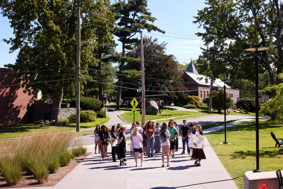 a group of people walking down a sidewalk next to a park and a street sign with a building in the background