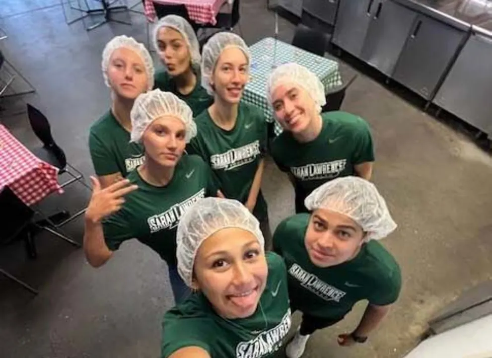 Group of seven people wearing green shirts and hairnets, posing indoors with checkered tablecloths in the background.