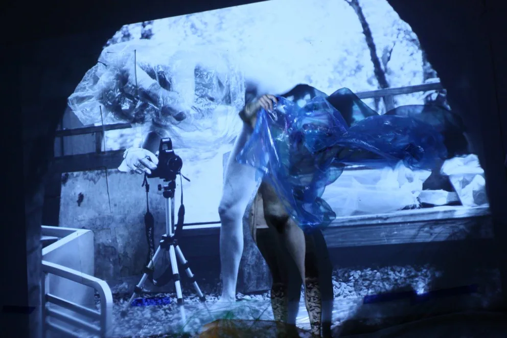 Abstract image showing a person wrapped in translucent material with a camera on a tripod, projected on a background of outdoor scenery.