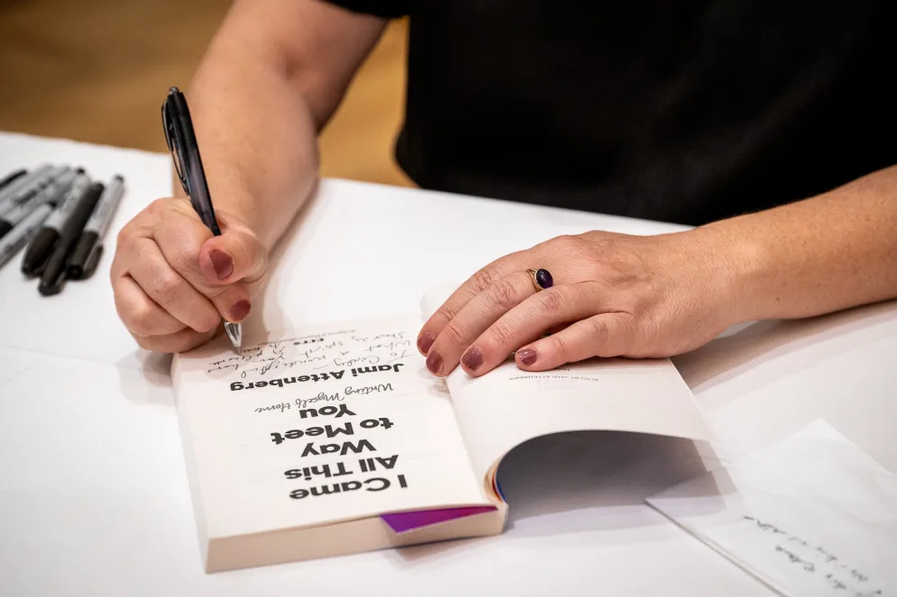 a person writing on a book with a pen and a notebook on a table next to a pair of gloves