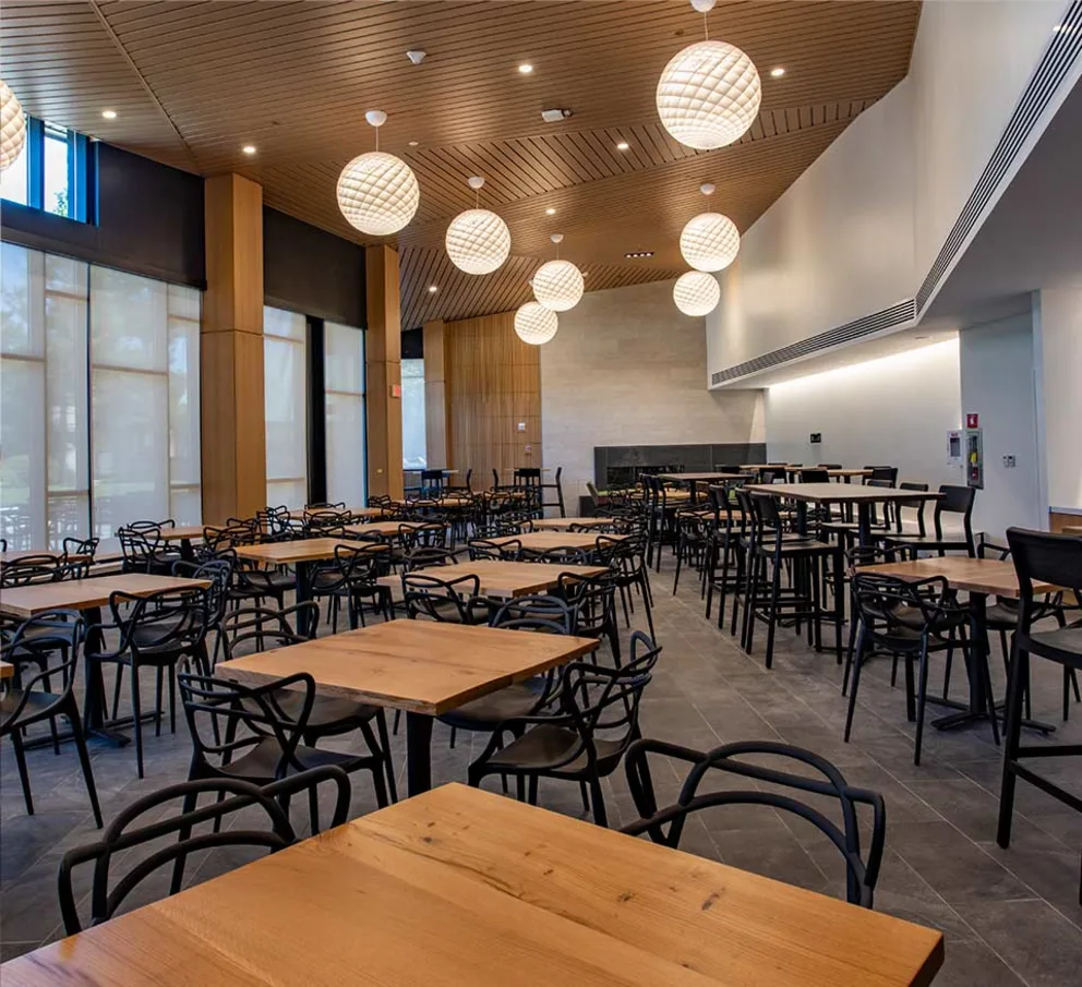 A modern cafeteria with wooden tables and black chairs, featuring globe lights on the ceiling and large windows with natural light.