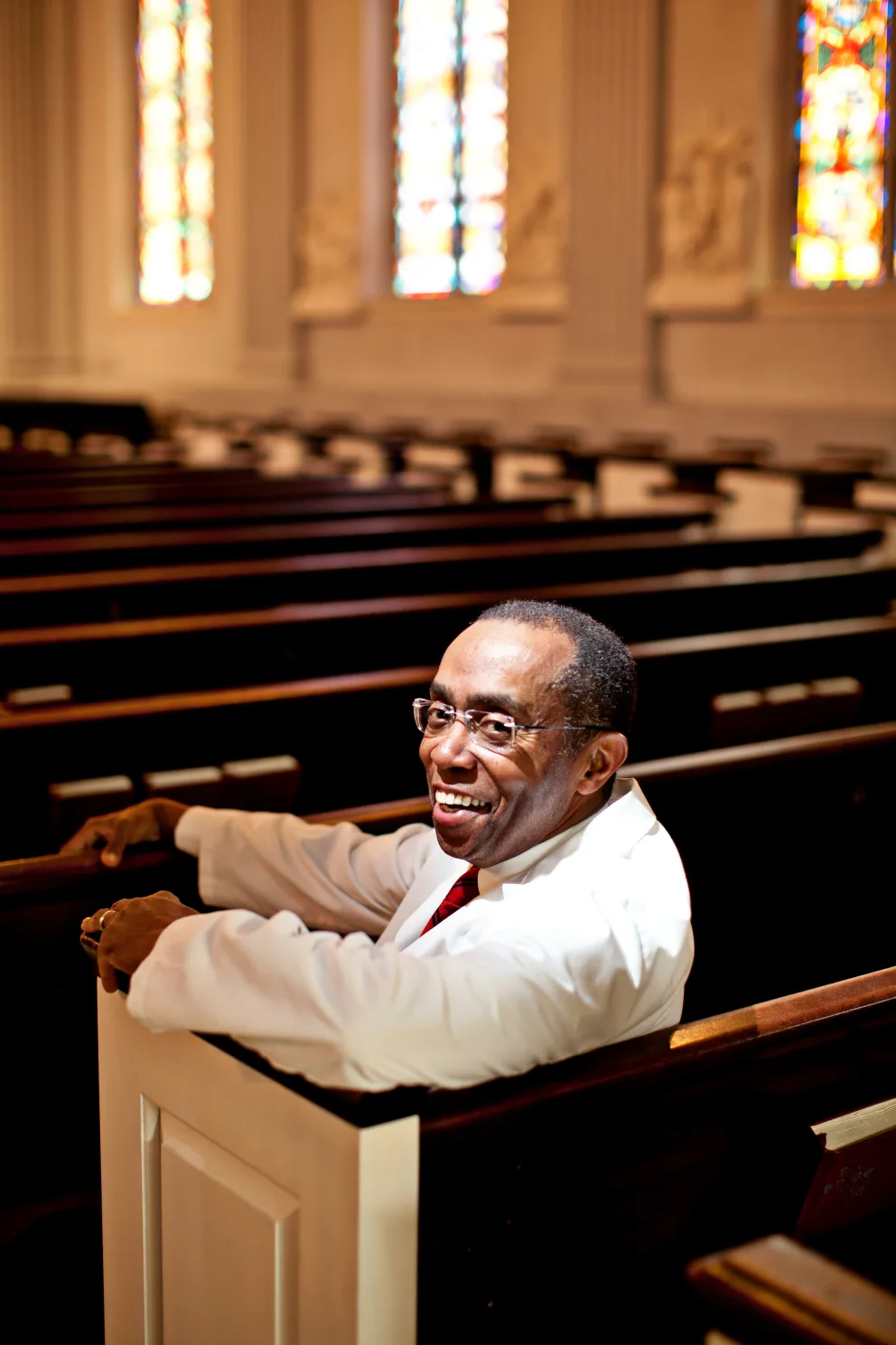 Person smiling and sitting in a church pew, surrounded by wooden benches and colorful stained glass windows.