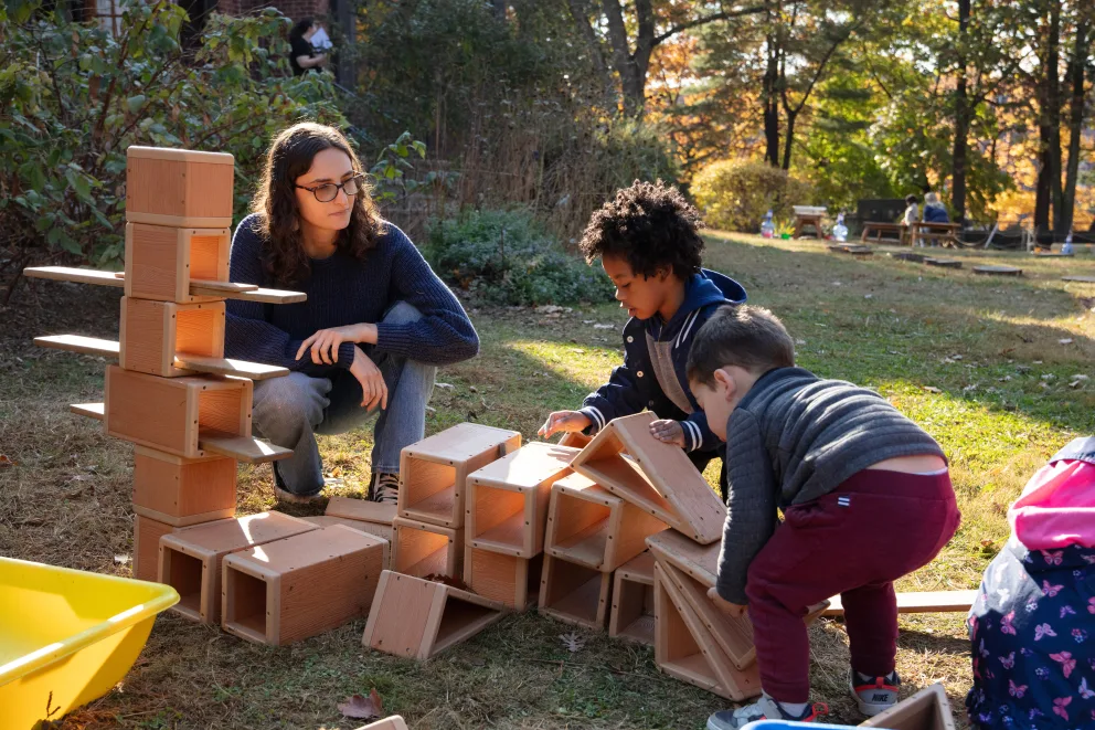 Person with children building a tall structure from wooden blocks on grass, surrounded by trees in a park setting.
