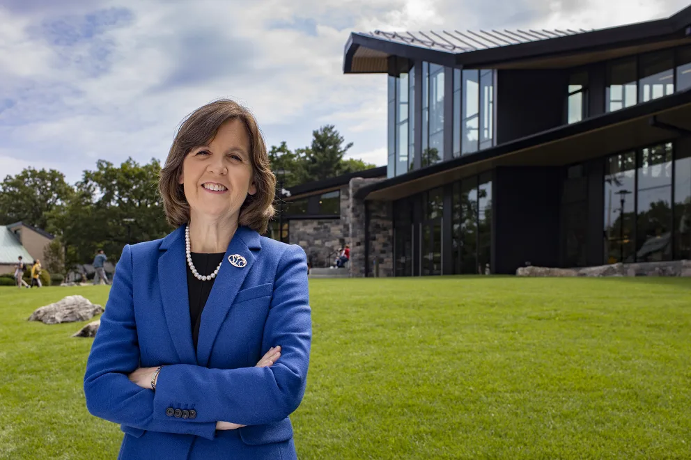 Person in a blue suit stands smiling with arms crossed on a grassy lawn, in front of a modern building with large windows.