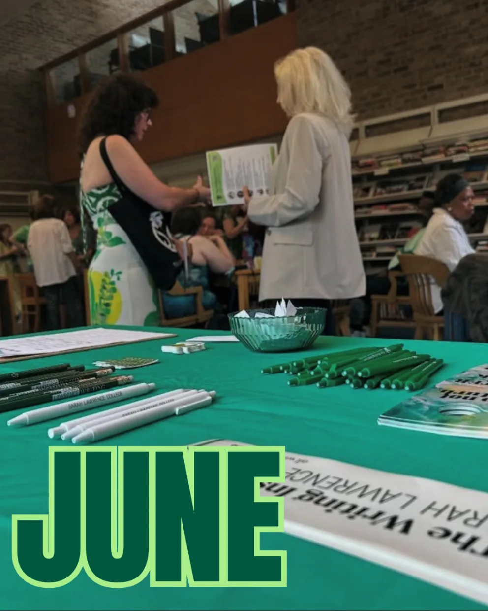 Tables with green pens and papers are in a busy library. Two people stand talking. The word "JUNE" is prominently displayed in green.