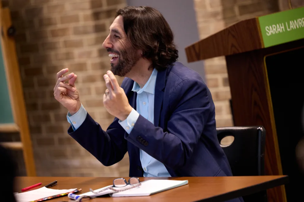 Person with long hair and beard smiling and gesturing during a talk at a table with notes and glasses, near a lectern with Sarah Lawrence label.
