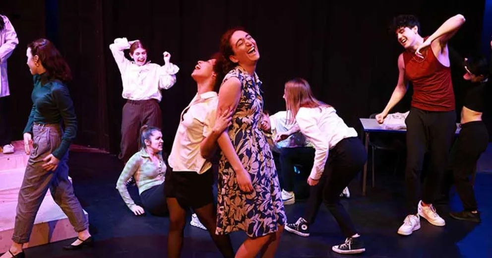 Performers on stage, energetically dancing and laughing. Background is dark, creating a focus on the dynamic expressions and movements.