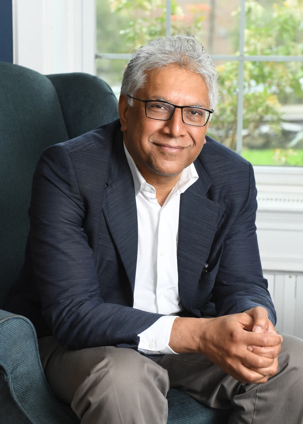 Person with gray hair and glasses, wearing a dark blazer and white shirt, seated on a chair indoors, with a window view in the background.