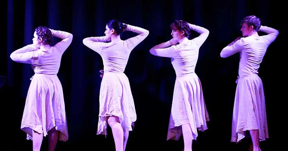 Four dancers in light dresses stand on stage with their hands on their heads, backs to the audience, under purple lighting.